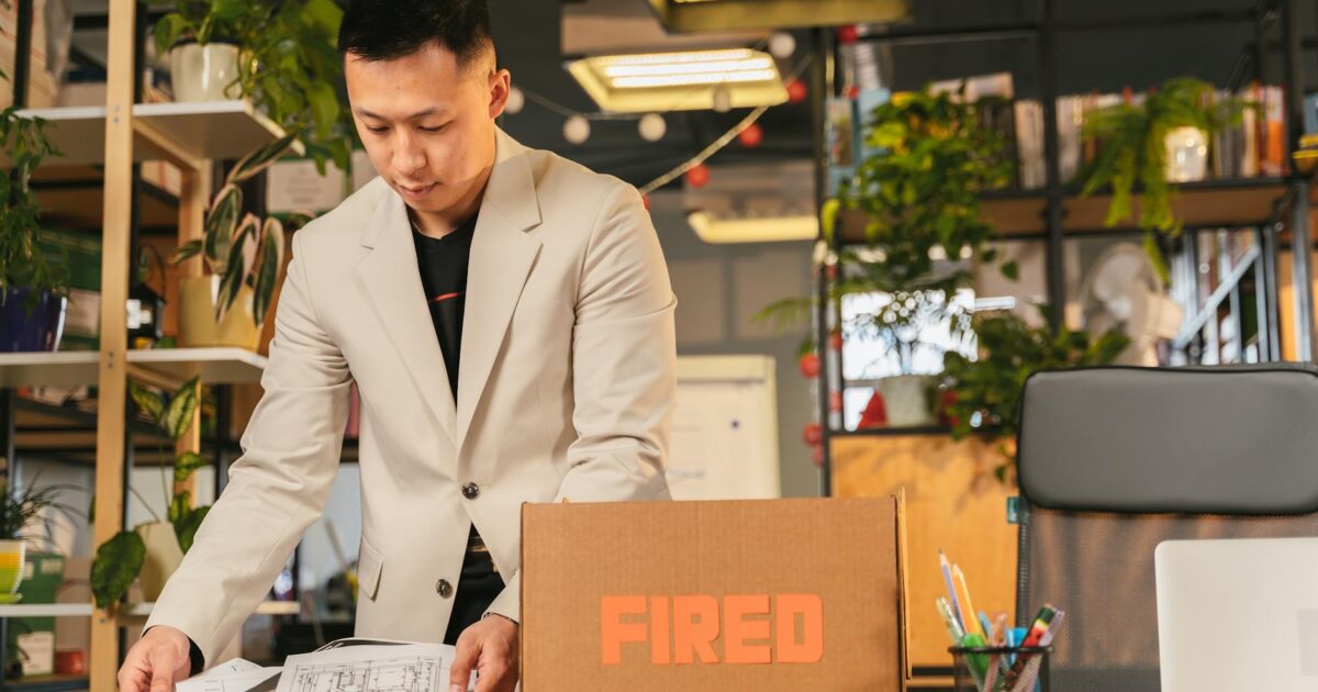 An employee packing documents into a box labeled 'FIRED' in an office environment.