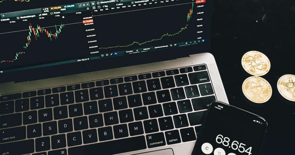 A laptop with trading charts, smartphone calculator, and bitcoin coins depicting cryptocurrency trading.